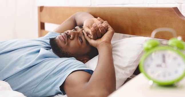 Black man laying in bed with green alarm clock.