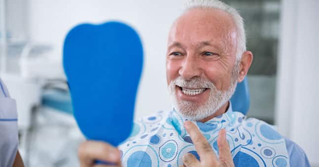 Older man smiling with mirror looking at new dental implants.